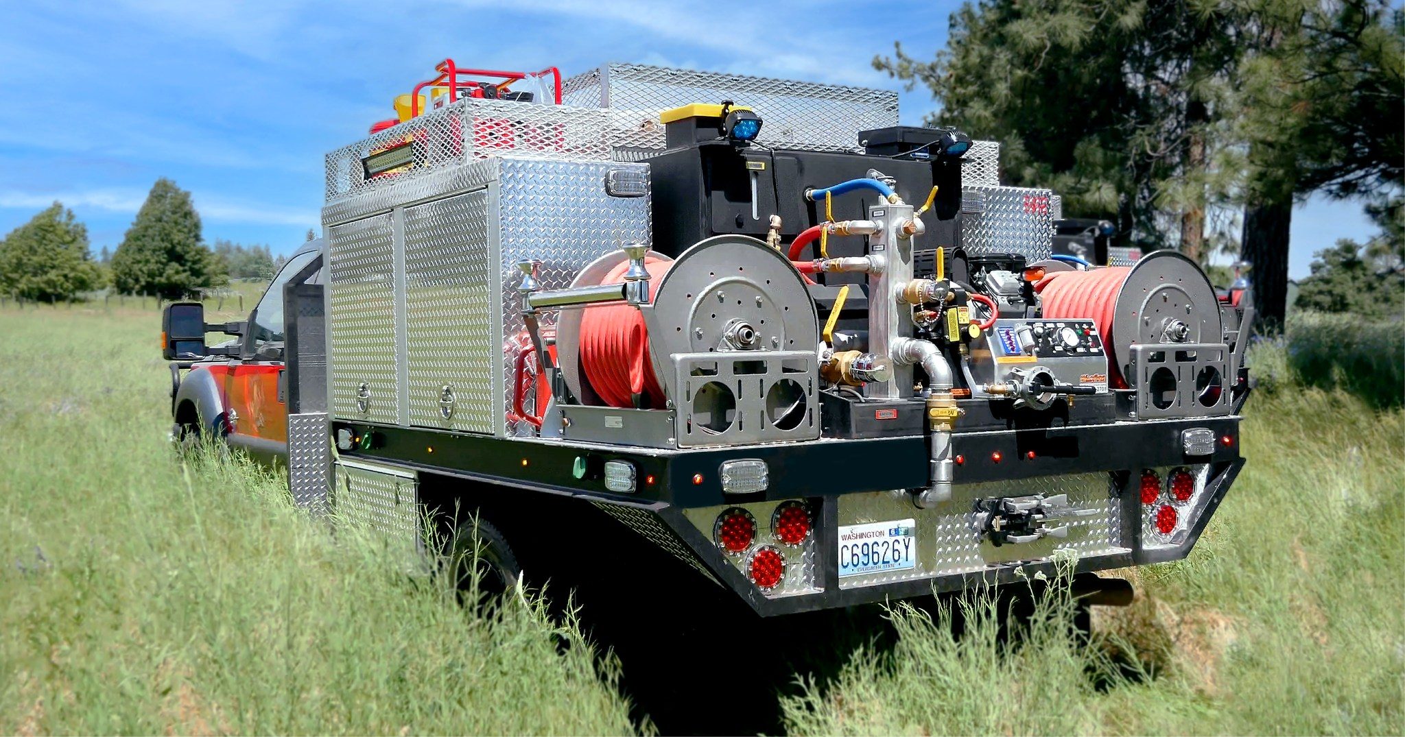 A custom-built Global Wildfire wildland fire rig on a flatbed truck with dual hose reels, pump panel, and diamond-plate compartments, parked in a grassy field